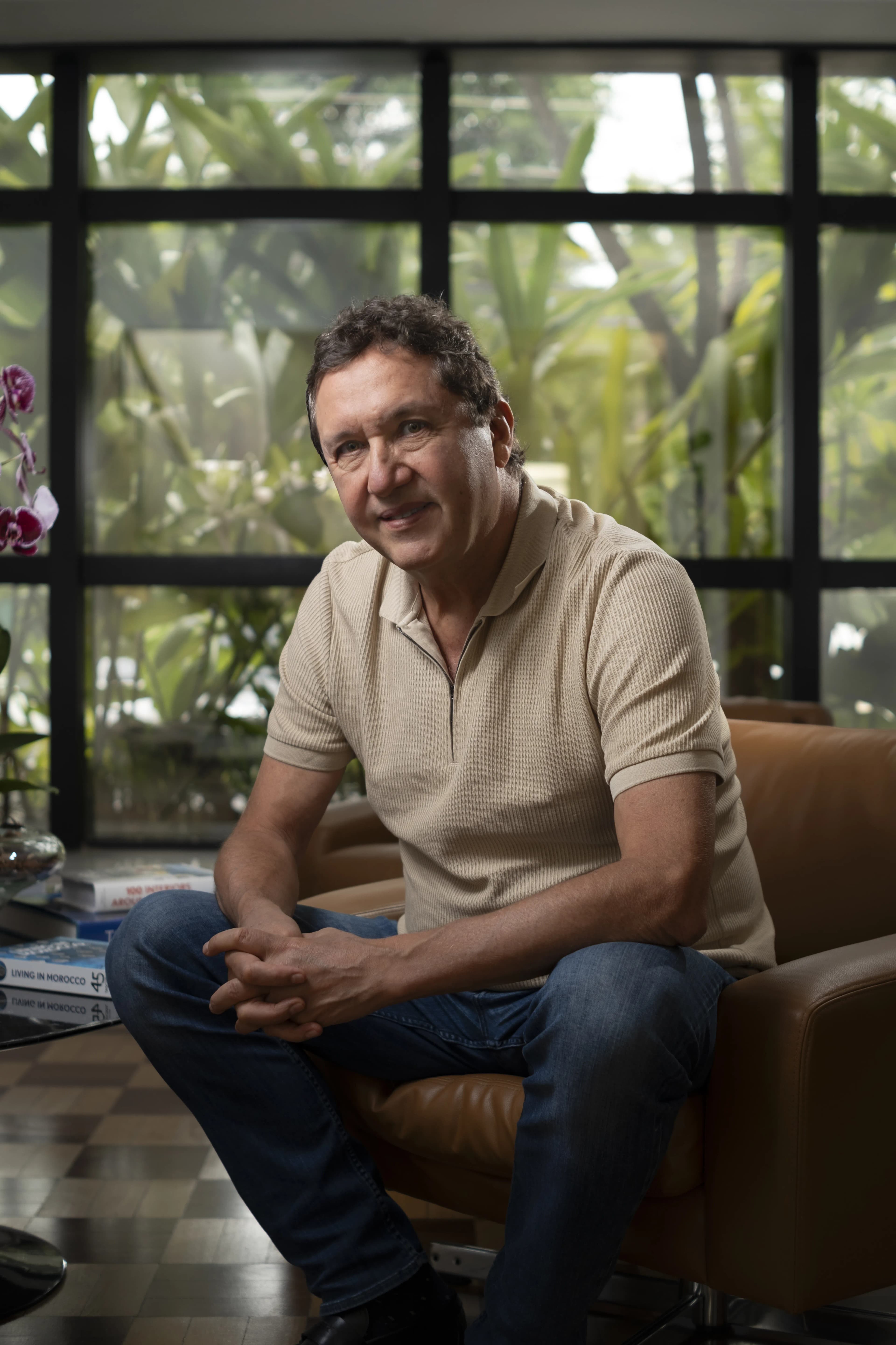 Portrait of Amir Makansi seated indoors in front of large windows and tropical greenery