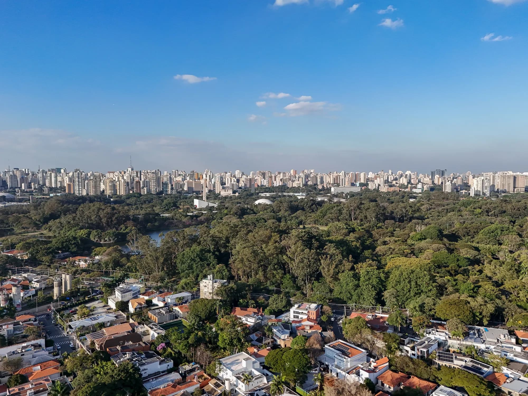 Aerial view of a sprawling city skyline beyond Ibirapuera park, with houses in the foreground under a bright blue sky