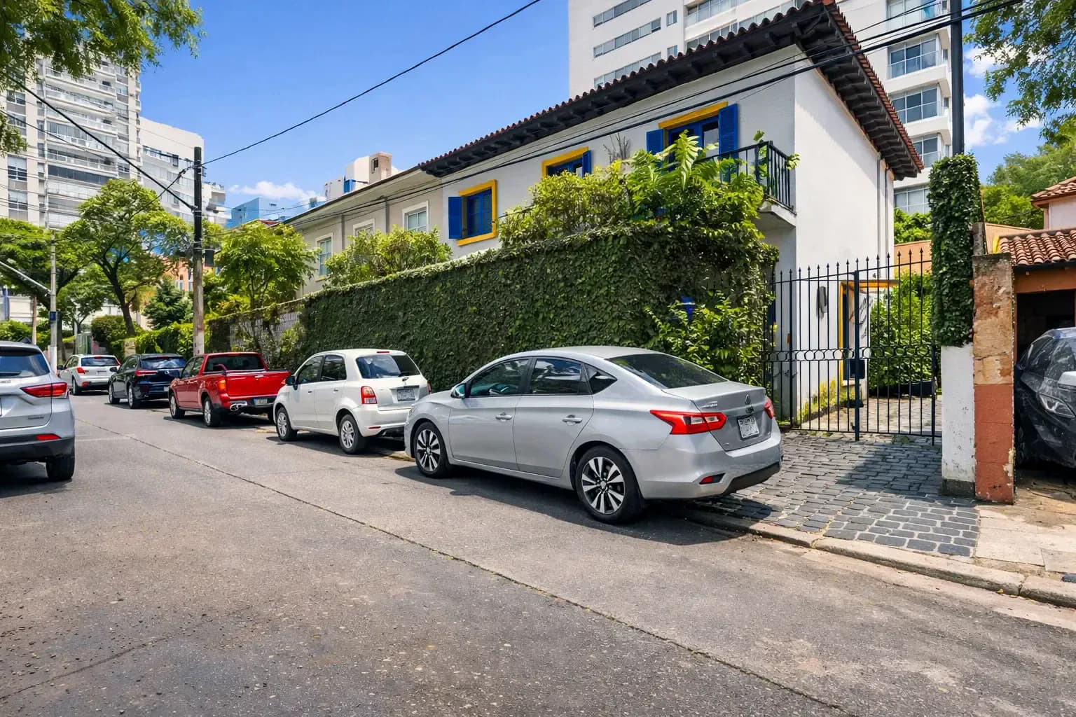 Residential street with parked cars beside an ivy-covered wall and white colonial-style house